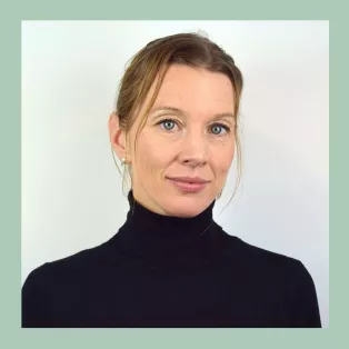 Portrait of Caroline Delgado who is looking into the camera with a gentle smile, wearing a black turtleneck. Photo.